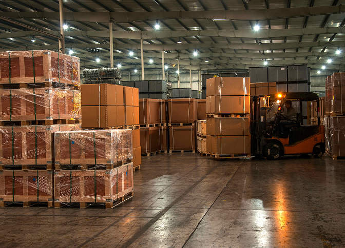 Forklift moving pallets of bulk shipping boxes in a large warehouse.